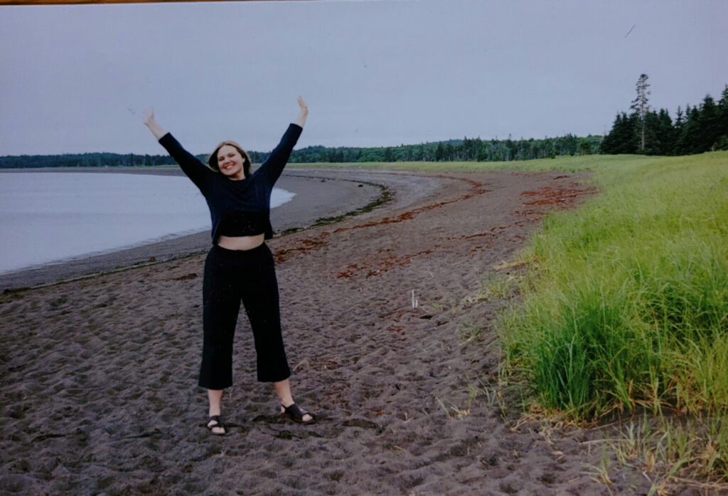 Person standing on a sandy beach with arms raised, near grassy dunes and calm water at Herring Cove.