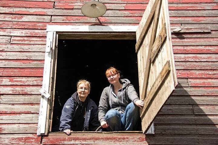 Two people sitting in an open barn loft doorway on a weathered red‑wood exterior.