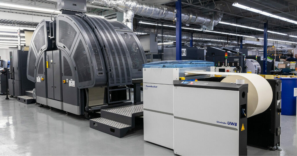 Inkjet web press running in a production facility, with large paper rolls feeding into the press and surrounding equipment visible on the shop floor.