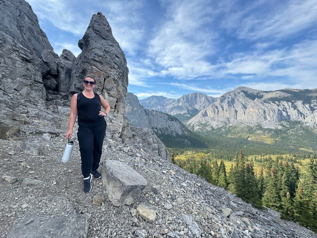 Person standing on a rocky mountain trail holding a water bottle, with dramatic peaks and forested valley behind.
