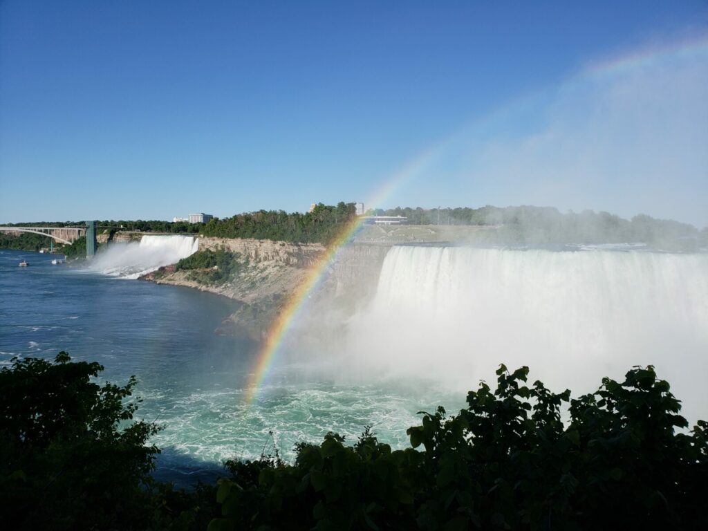 Rainbow arching over Niagara Falls with mist rising and bright blue sky above.