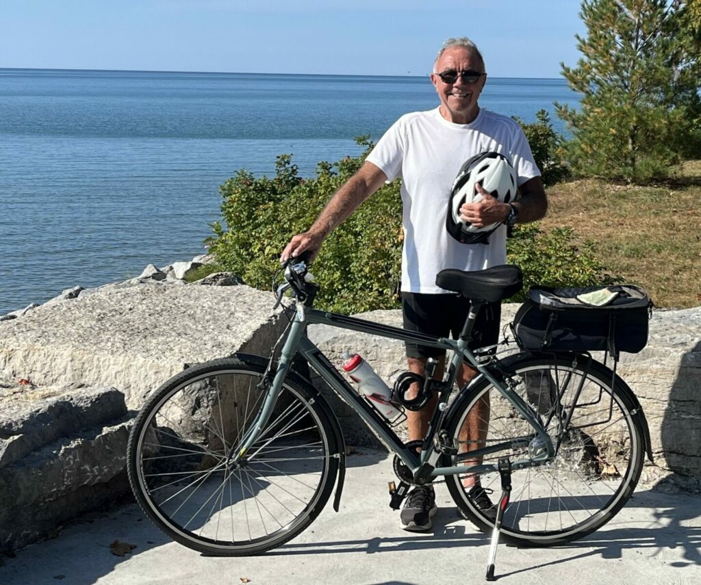 Person standing with a bicycle on a lakeside path, holding a helmet with Lake Huron in the background.