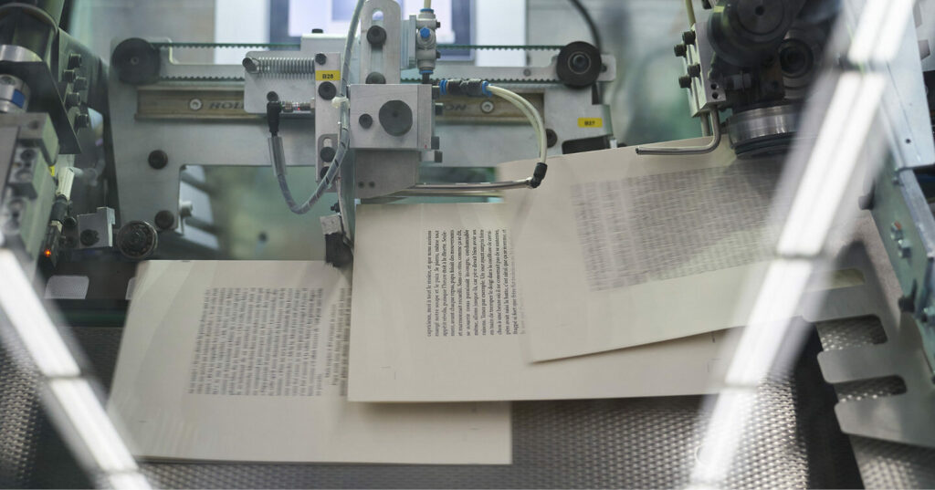 Close-up view of sheets moving through an automated book‑binding machine, with mechanical arms and rollers guiding the printed pages.