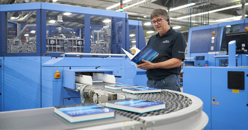 A worker stands beside a blue book‑encasing machine, examining a finished hardcover book as additional copies move along the conveyor system.