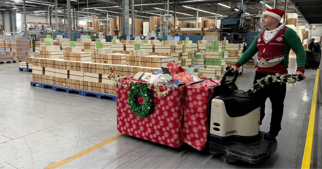 A decorated cart covered in red gift‑wrap and tinsel being pulled through a production floor, surrounded by stacks of book signatures.