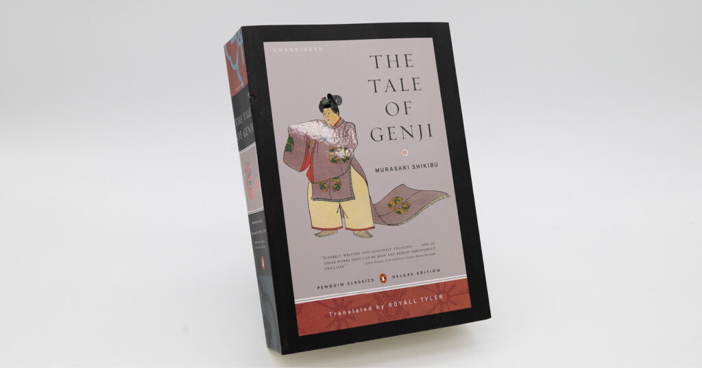 Front cover of ‘The Tale of Genji’ by Murasaki Shikibu, deluxe edition, featuring a traditional illustrated figure against a soft grey background, photographed standing upright on a white surface.
