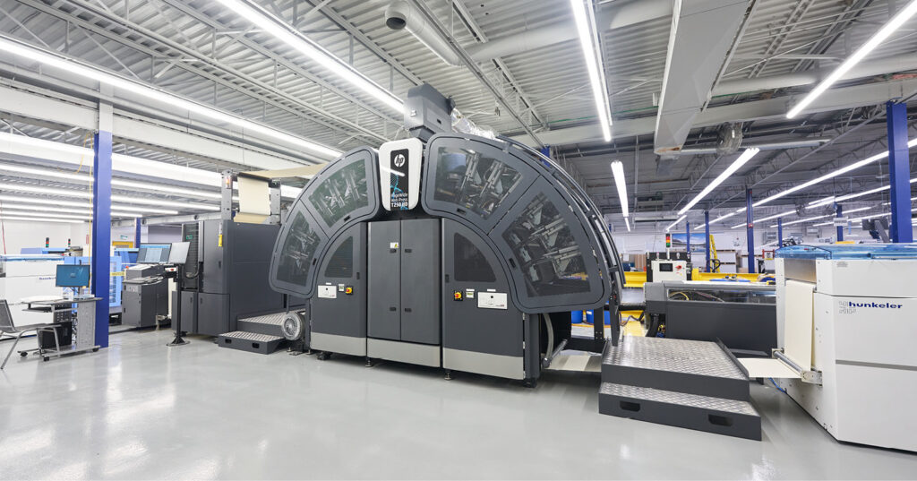 Large HP inkjet printing press in a production facility, featuring a curved high‑capacity print engine and connected paper‑handling units under bright overhead lighting.