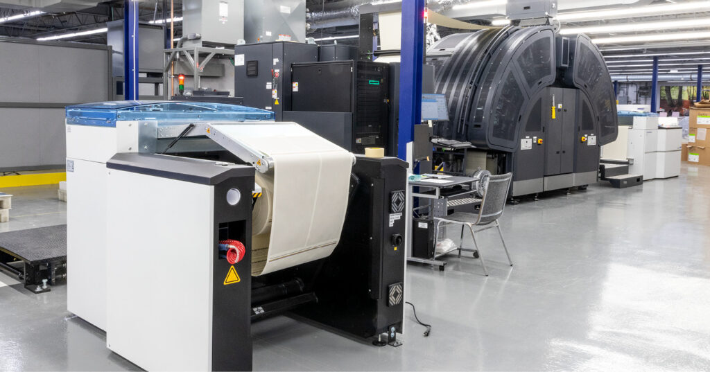Large inkjet printing press in a production facility, with a roll-fed paper unit in the foreground and a curved high‑capacity print engine in the background.