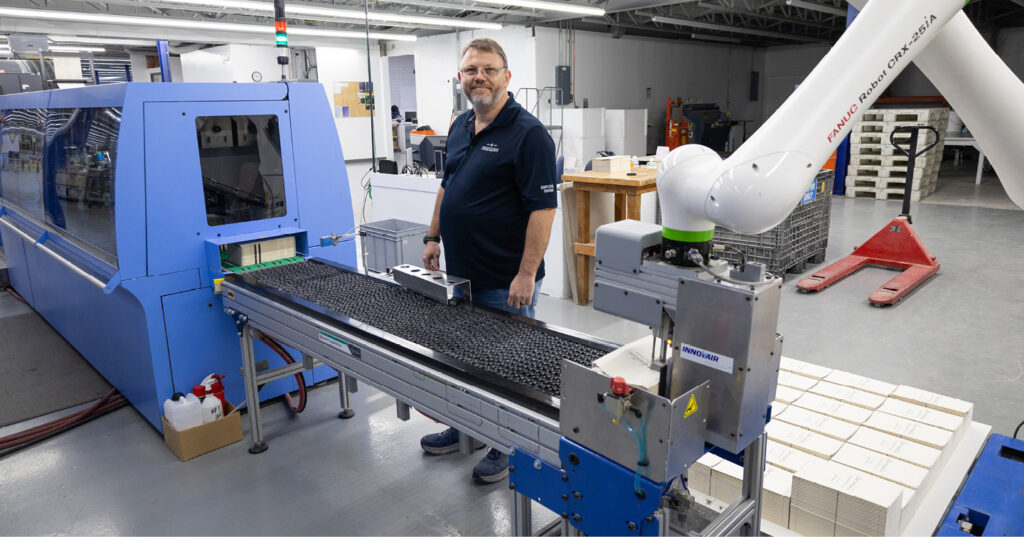 Worker operating a robotic arm beside a conveyor system attached to a large blue industrial press inside a manufacturing facility.