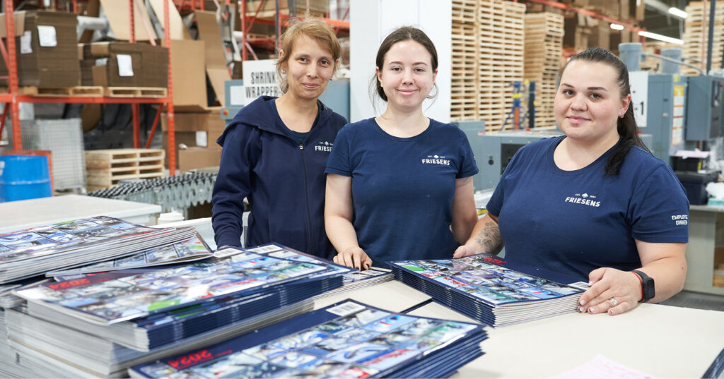 Three Friesens employees standing at a work table in the production floor, with stacks of printed materials laid out in front of them.