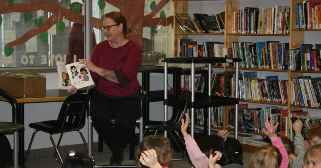 An adult seated in a library reading a picture book to a group of children who are sitting on the floor with hands raised.