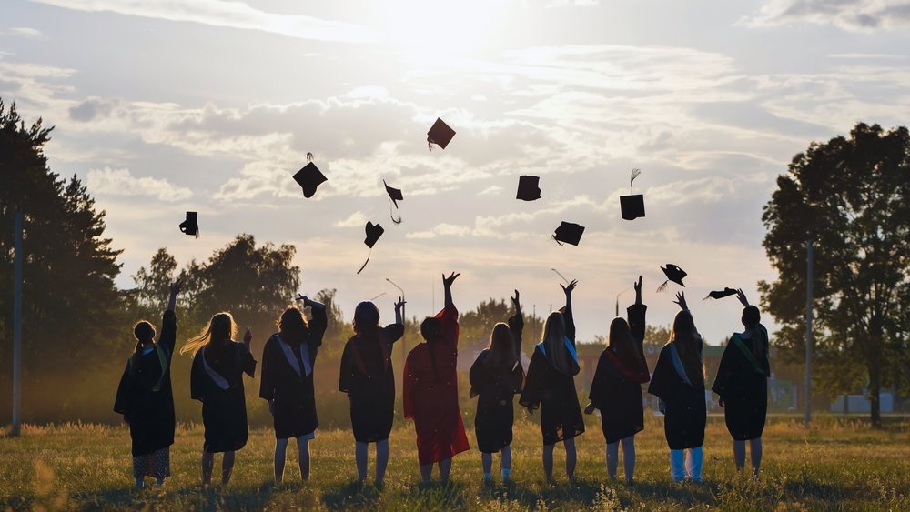 Group of graduates in caps and gowns standing outdoors at sunset, throwing their graduation caps into the air.