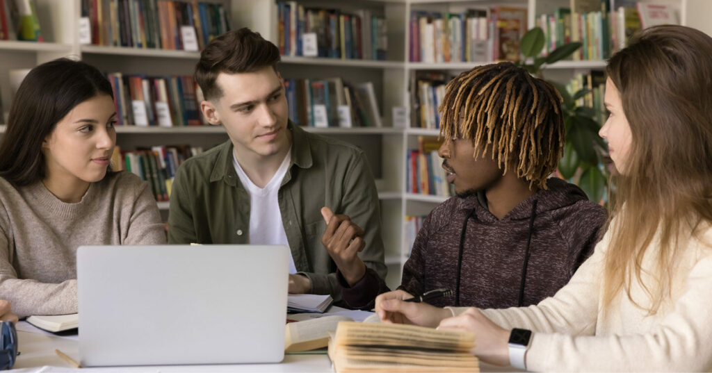A group of students sitting together at a table in a library, talking and working with books and a laptop.