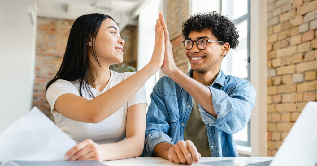 Two people sitting at a table, smiling and giving each other a high‑five while working with papers and a laptop.