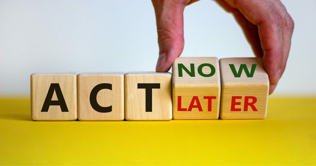 A hand rearranging wooden blocks to change the message from ‘Act Later’ to ‘Act Now’ on a yellow surface.
