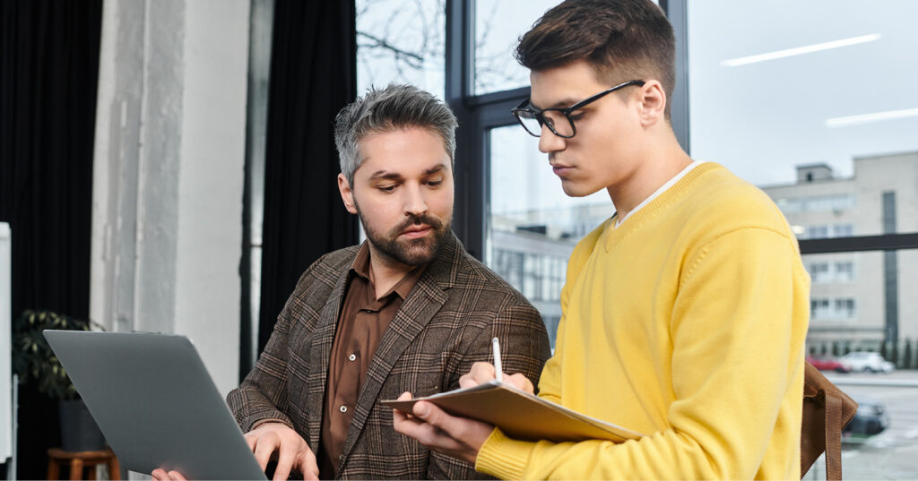 Two coworkers discussing notes beside a laptop in a bright office with large windows.