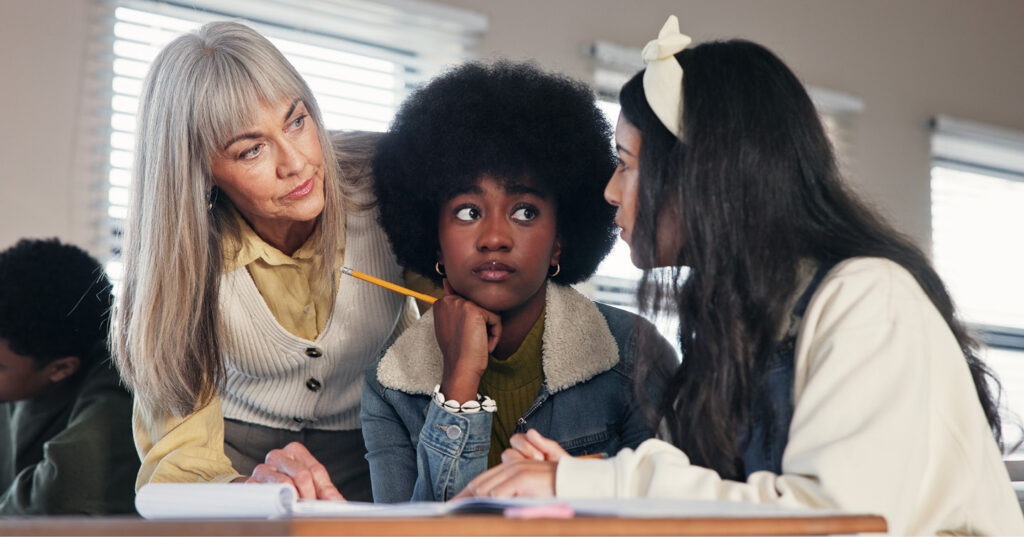 Teacher leaning over a desk, helping students review notes and discuss work in a bright classroom setting.