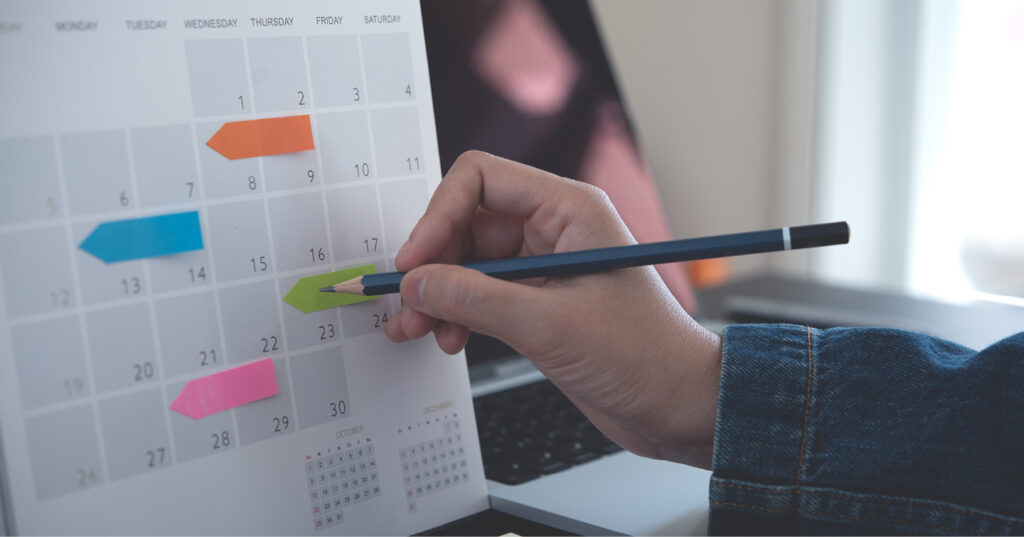 Hand holding a pencil, marking a date on a desk calendar with colorful sticky notes and a laptop in the background.