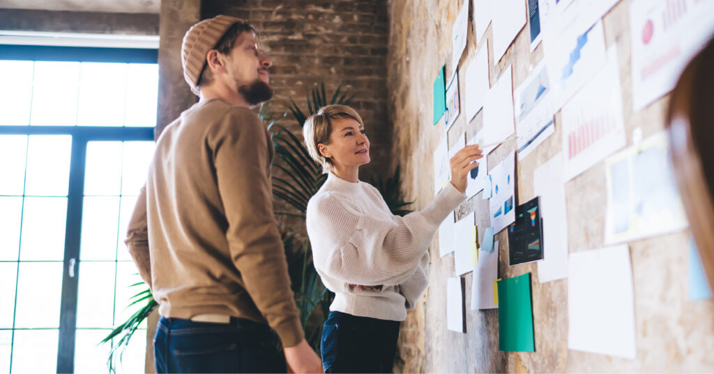 Two coworkers reviewing notes and charts pinned to a wall in a creative office during a planning session.