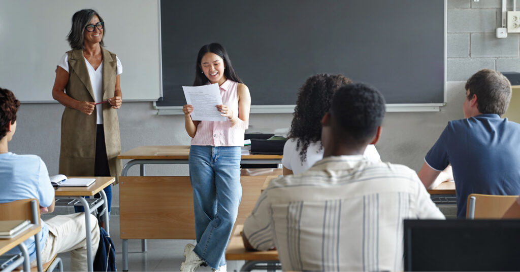 Student presents a paper at the front of a classroom while a teacher stands nearby and classmates listen.