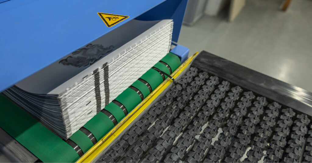 Stack of printed sheets moving on green conveyor rollers toward a finishing or processing station.