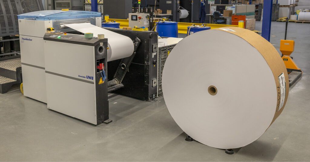Large roll of paper mounted on an industrial unwinder machine inside a print production facility.