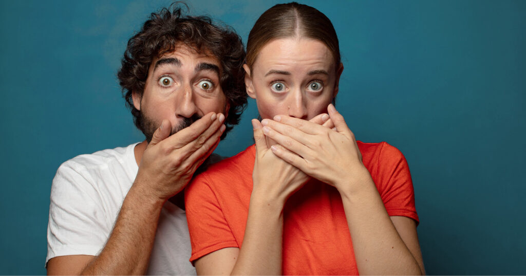 Two people standing close together, hands covering their mouths, against a plain blue background.