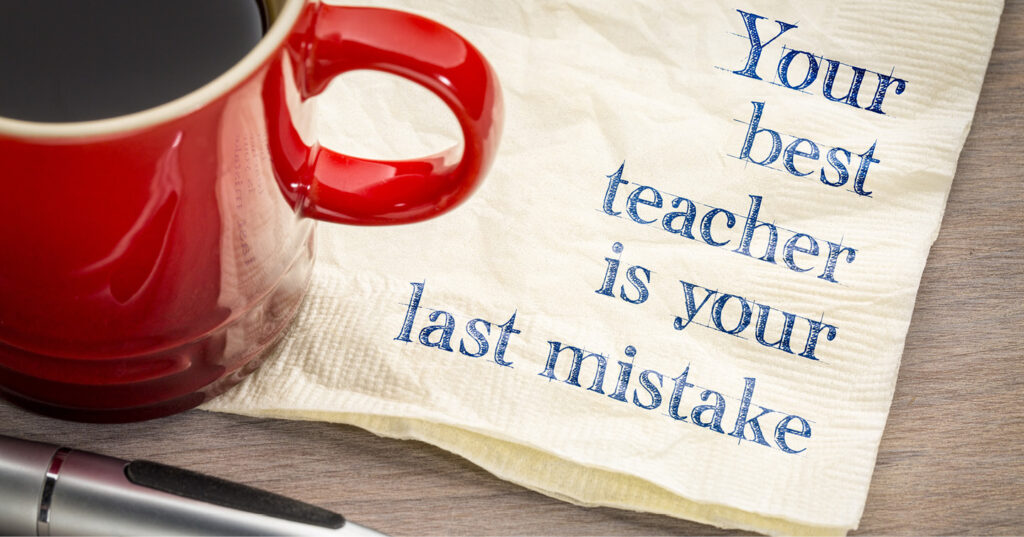 Red coffee mug beside a napkin printed with the quote “Your best teacher is your last mistake” on a desk.