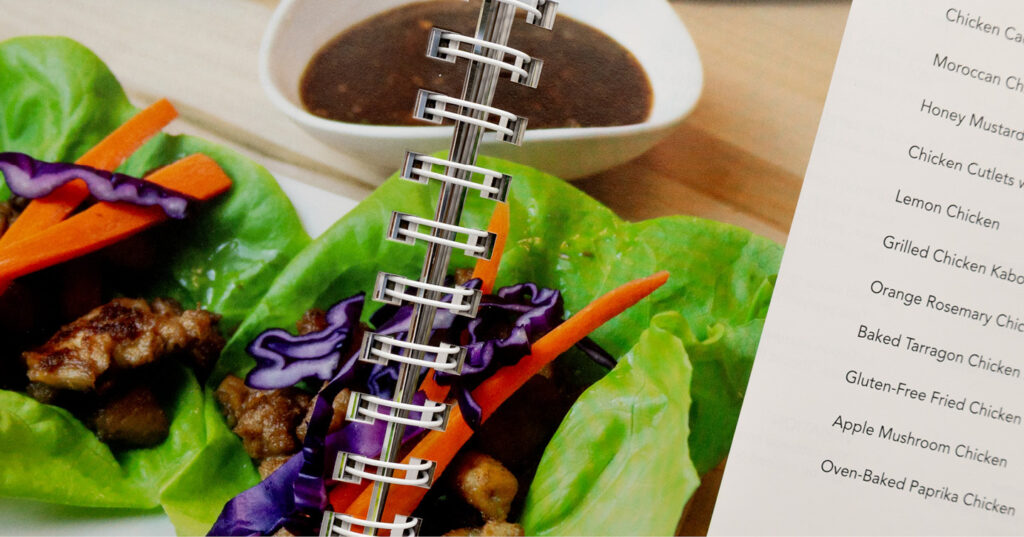 Close-up of a spiral-bound cookbook showing chicken lettuce wraps with sauce and a list of chicken recipes.