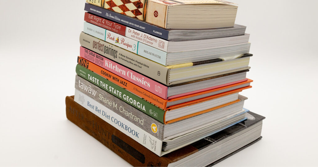 Tall stack of assorted cookbooks piled neatly on a white surface, showing colorful spines and varied thicknesses.