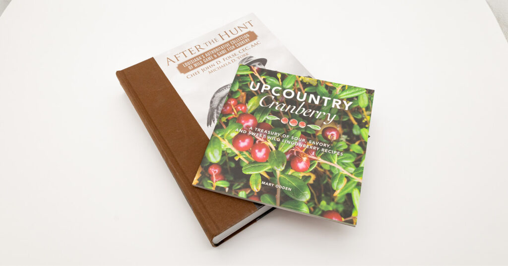 Two cookbooks stacked on a white surface, featuring a cranberry-themed cover and a brown hardcover food book.