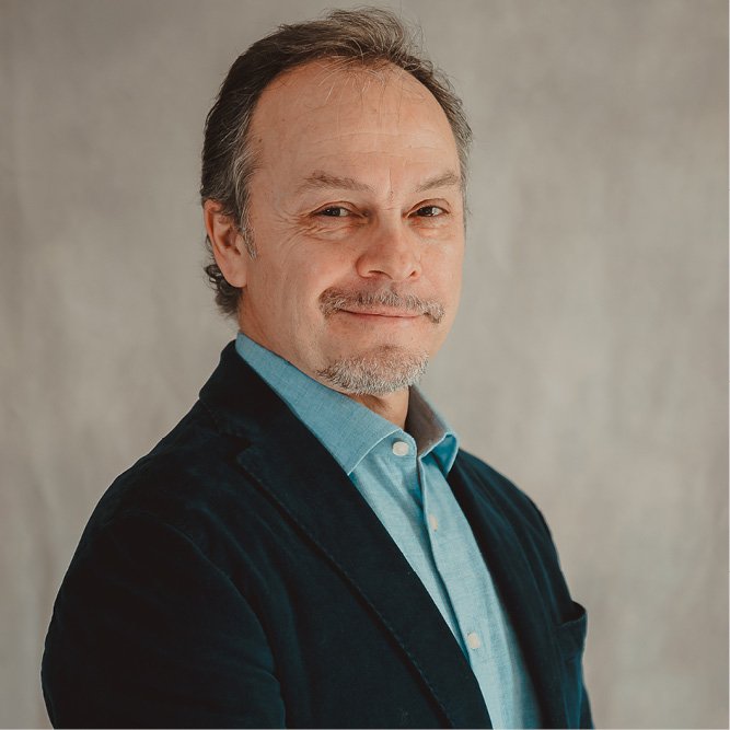 Studio portrait of a man wearing a blue shirt and dark jacket against a neutral background.