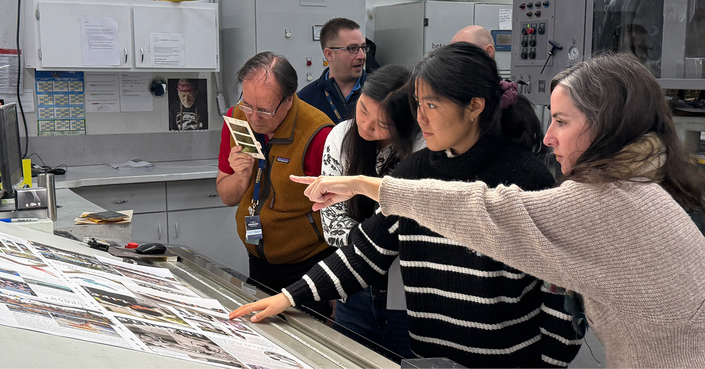 Team gathered around a press table, reviewing and pointing at printed pages in a production workspace.