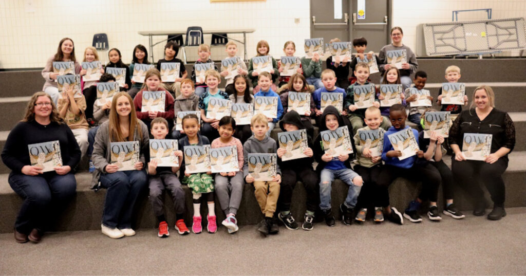 Group of Grade 2 students seated on risers holding copies of Owl during an I Love to Read Month school visit.