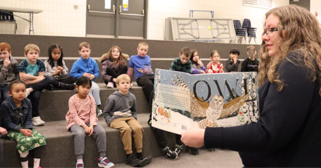 Adult holding an open copy of Owl while reading aloud to Grade 2 students seated on risers during a school visit.