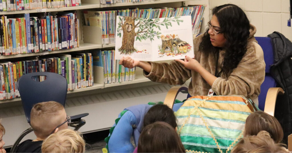 Adult reading Owl aloud to a group of young students in a school library, holding the open book to show illustrations.