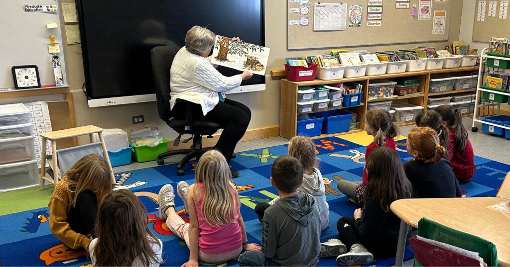 Adult reading Owl aloud to seated Grade 2 students in a classroom, holding the open book to show illustrations.