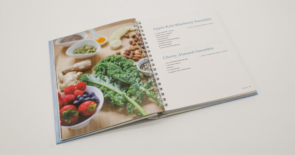Open spiral-bound cookbook showing smoothie recipes alongside fresh ingredients like berries, kale, nuts, and ginger.