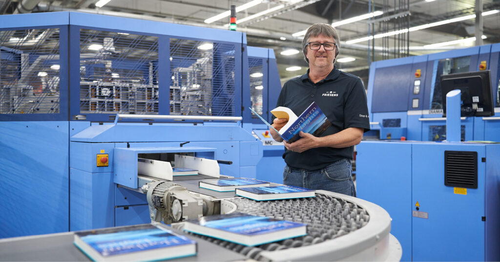 Employee-owner inspecting printed books beside automated finishing equipment on the production floor at Friesens.