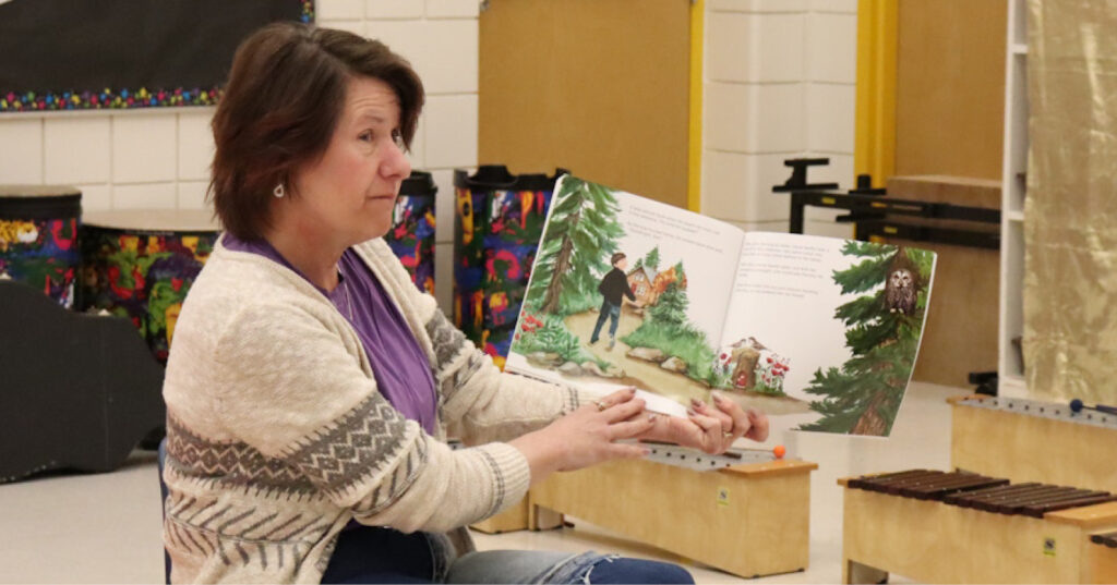 Adult holding an open copy of Owl, showing illustrated forest scenes while reading aloud during a school visit.