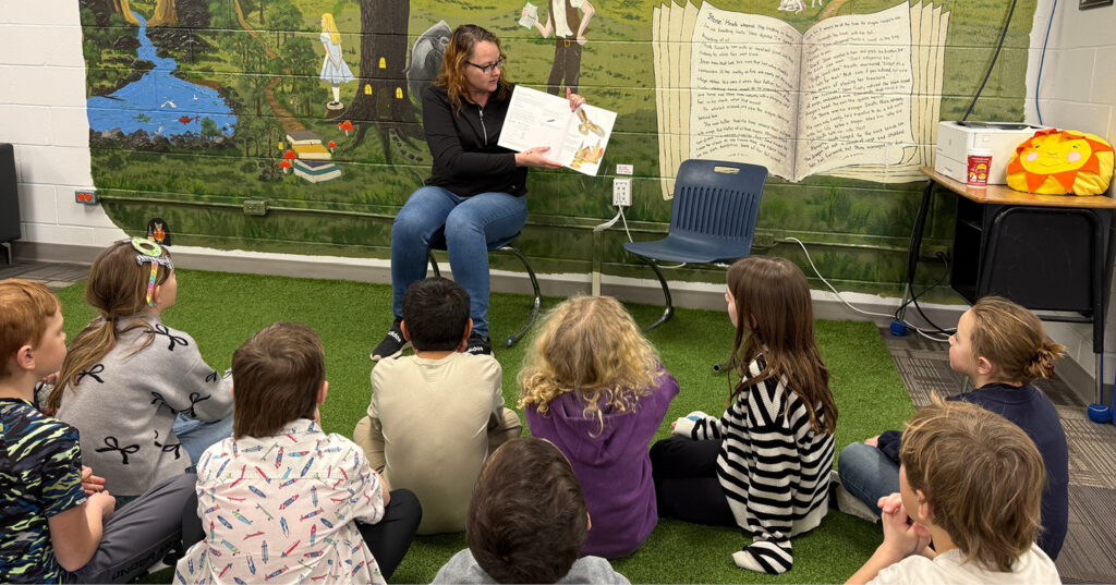 Adult reading Owl aloud to a group of Grade 2 students seated in a circle on classroom turf during a school visit.