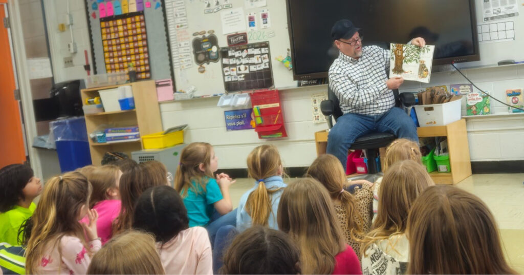 Adult reading Owl aloud to a group of Grade 2 students seated on the classroom floor during a school visit.