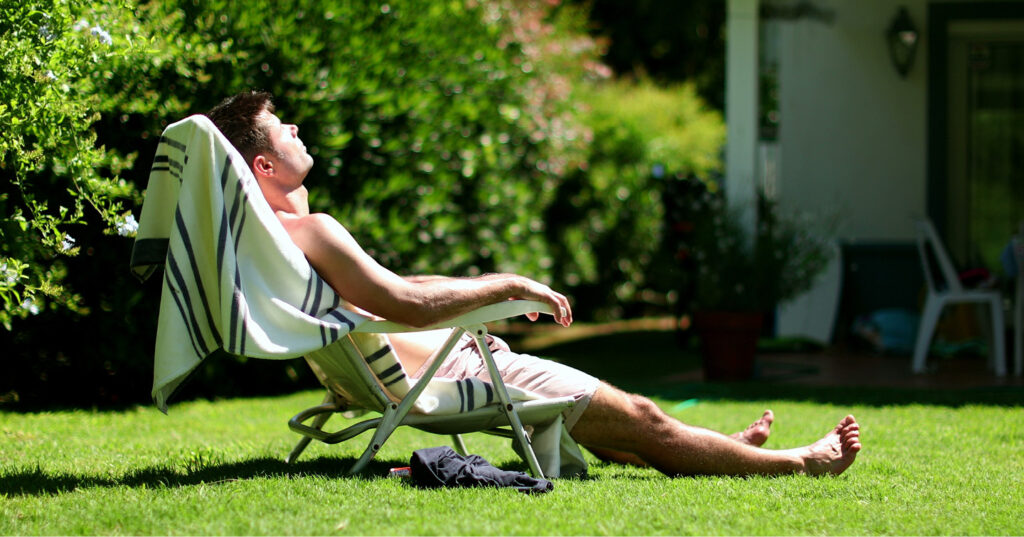 Person reclining on a lawn chair on green grass, enjoying sunlight in a quiet backyard garden.