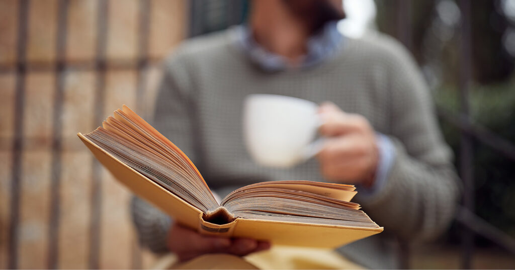 Person holding an open book while sipping from a white cup in a cozy, softly lit setting.