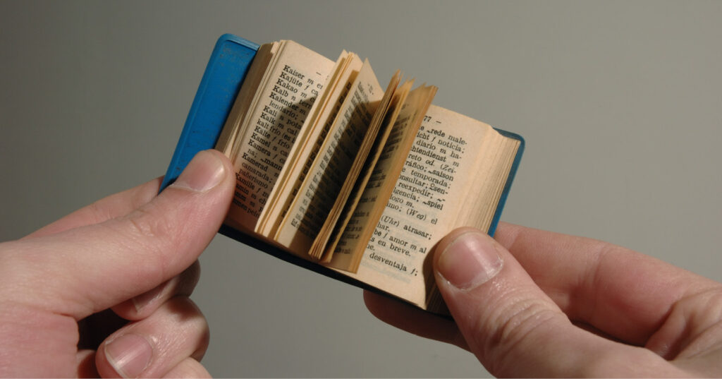 Close-up of hands holding a very small open book with thin pages and printed text.