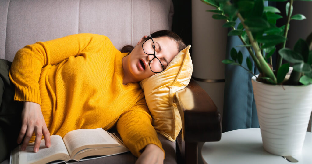 Person in a yellow sweater resting on an armchair with an open book, pillow, and houseplant nearby.
