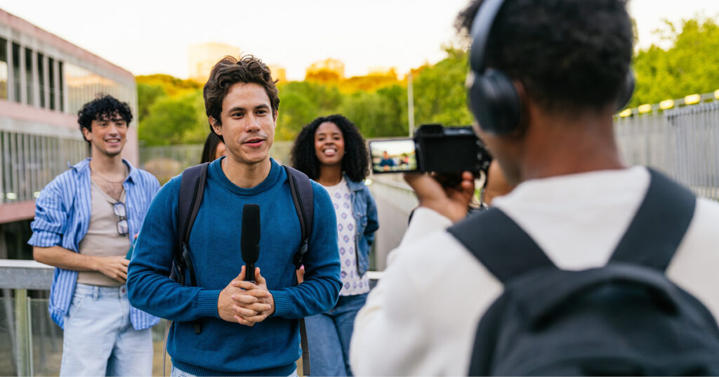 Student speaking into a microphone while being recorded by a peer with a camera, with classmates standing behind outdoors. Student