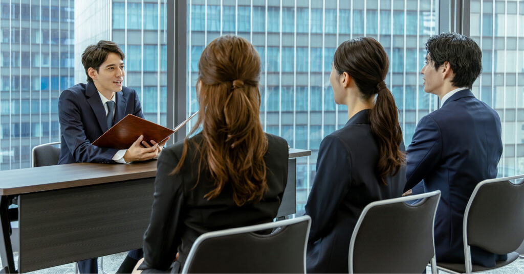 Job candidates seated at a table during an interview, listening to an interviewer holding a folder in a modern office setting.