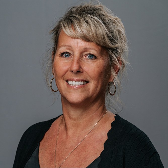 Person with gray hair wearing a black top and necklace, photographed against a neutral gray background.