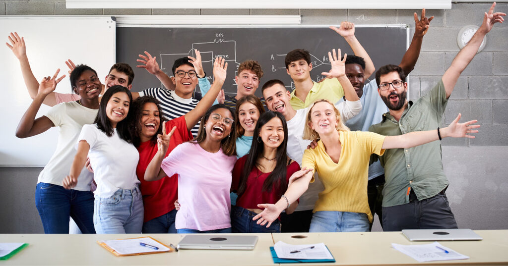 Group of students standing together in a classroom with raised arms, celebrating in front of desks and a chalkboard.